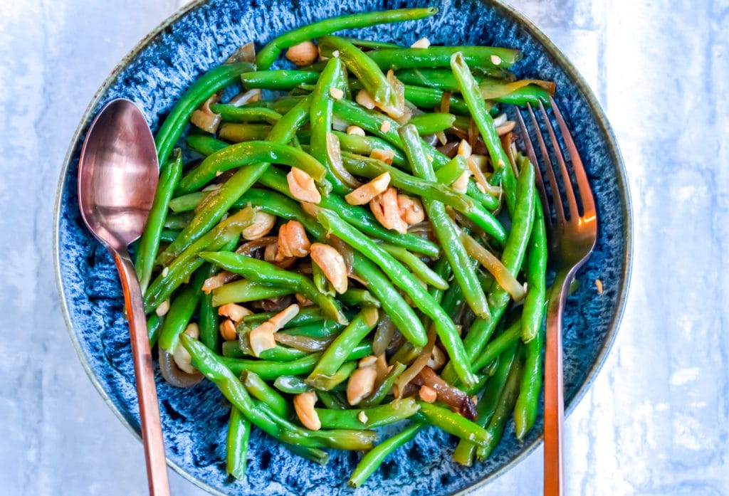 Green Beans with Caramelized Onions & Cashews  in a blue bowl with fork and spoon